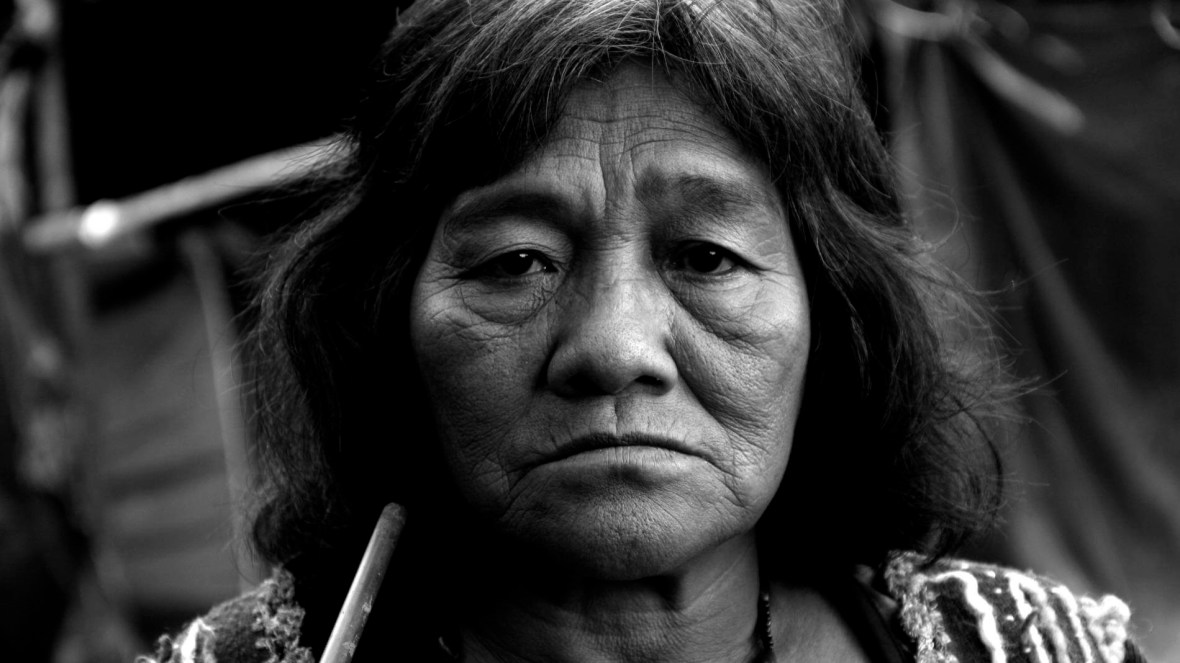 Mulher idosa Guarani-Mbyá olha séria para câmera enquanto segura um cachimbo